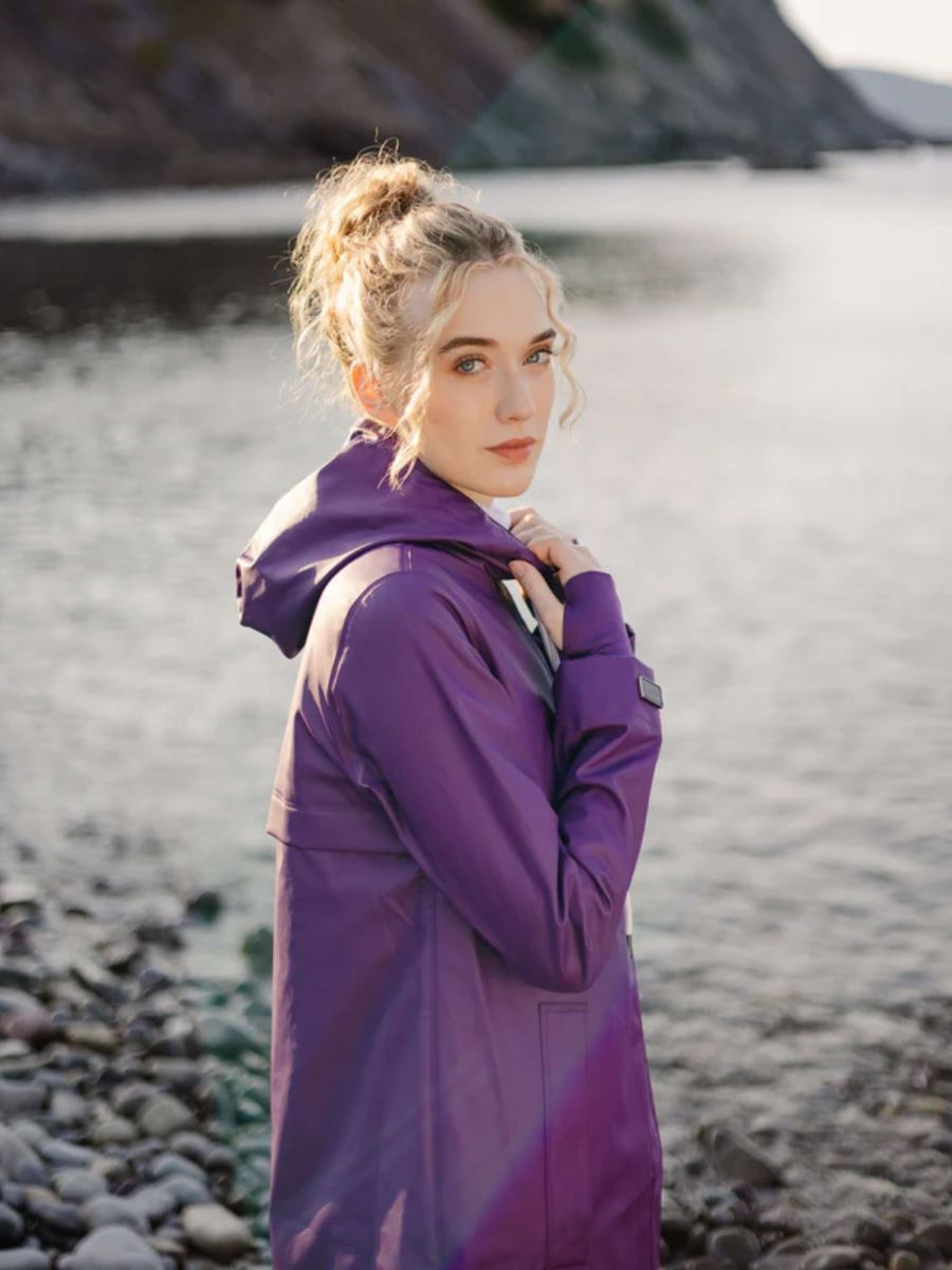 Individual wearing a purple waterproof coat standing by a rocky shoreline with water and cliffs in the background