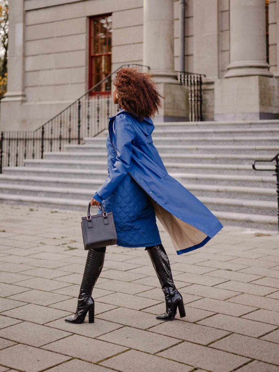 Person walking outside wearing a flowing blue trench coat, black knee-high boots, and holding a black handbag
