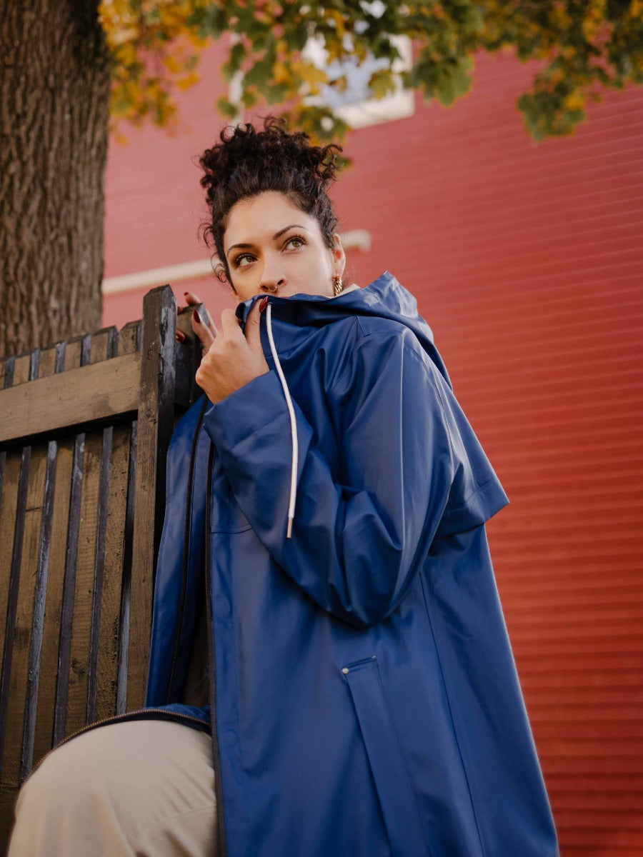 Person wearing a blue hooded raincoat outdoors near a wooden fence and tree with autumn leaves above