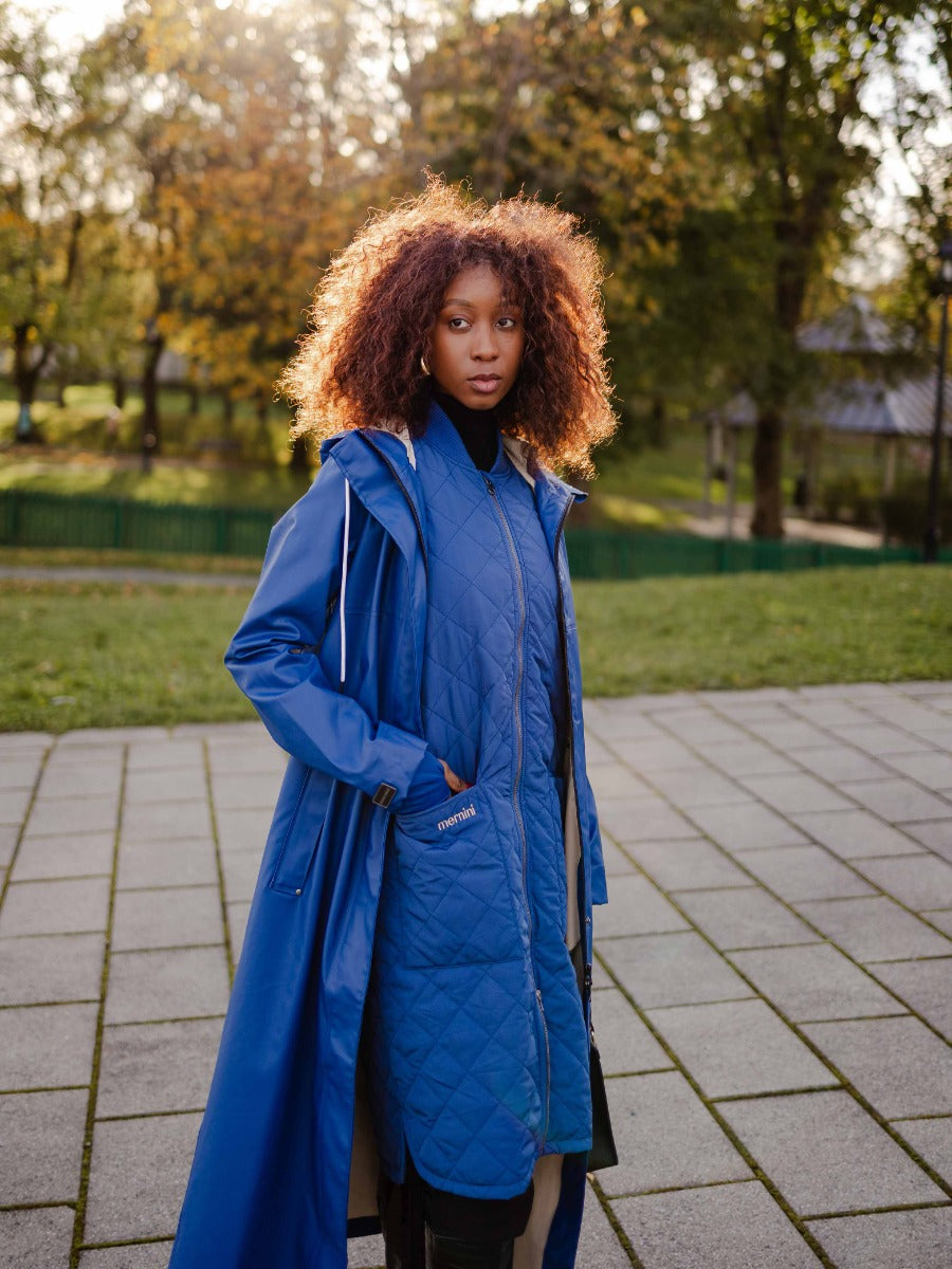 Person outdoors wearing a layered blue quilted jacket and long blue coat with autumn trees in the background