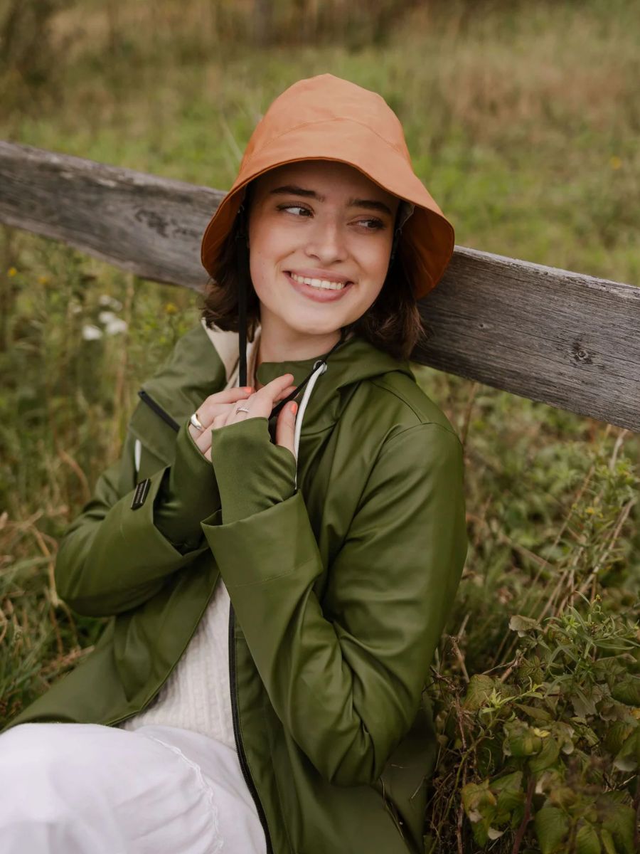 Smiling individual outdoors wearing a caramel-colored Mernini Waterproof Rain Hat by MERNINI/ROCK RAIN INC with green jacket.