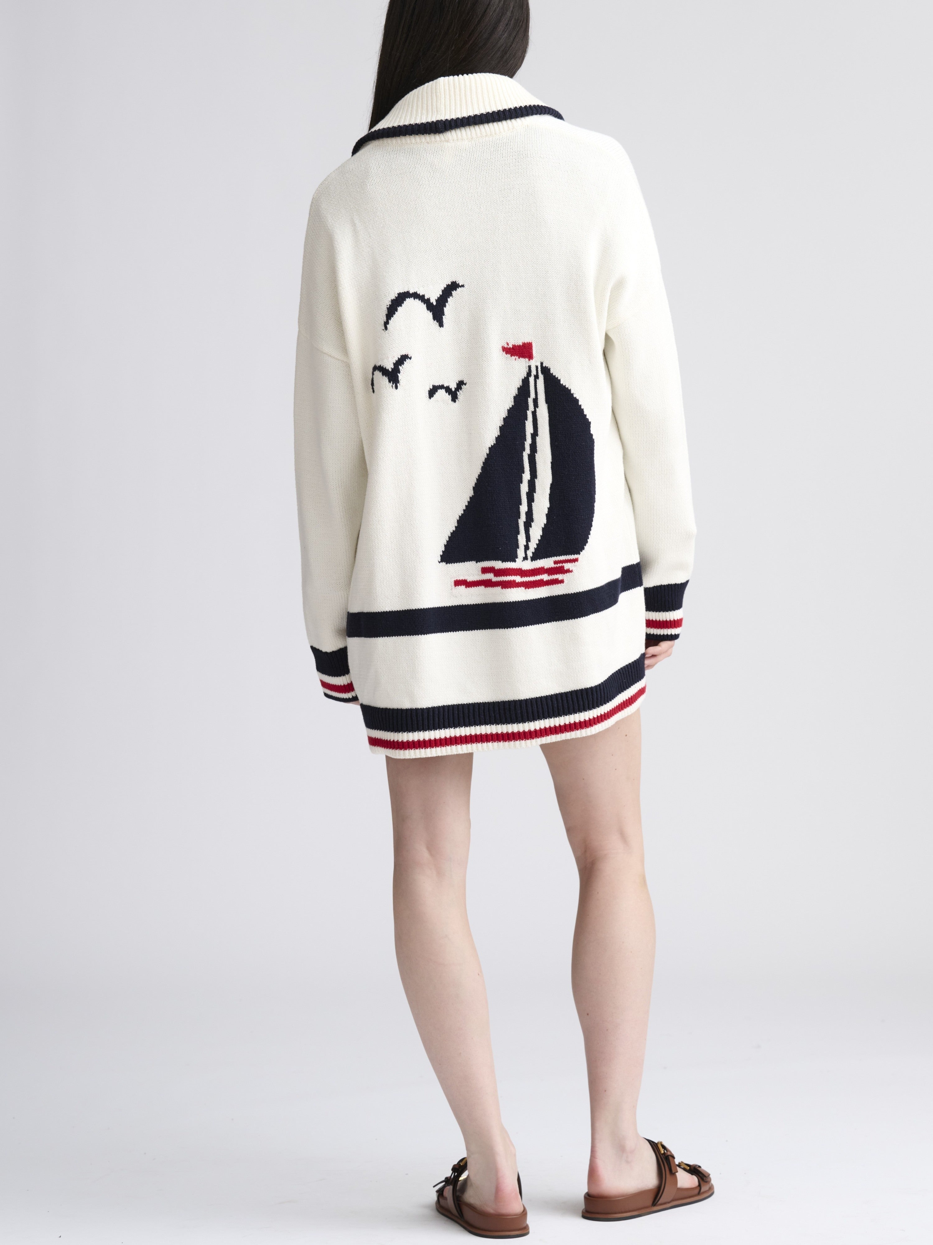 White knit sweater with navy and red sailboat and seagull patterns on the back, worn with sandals.