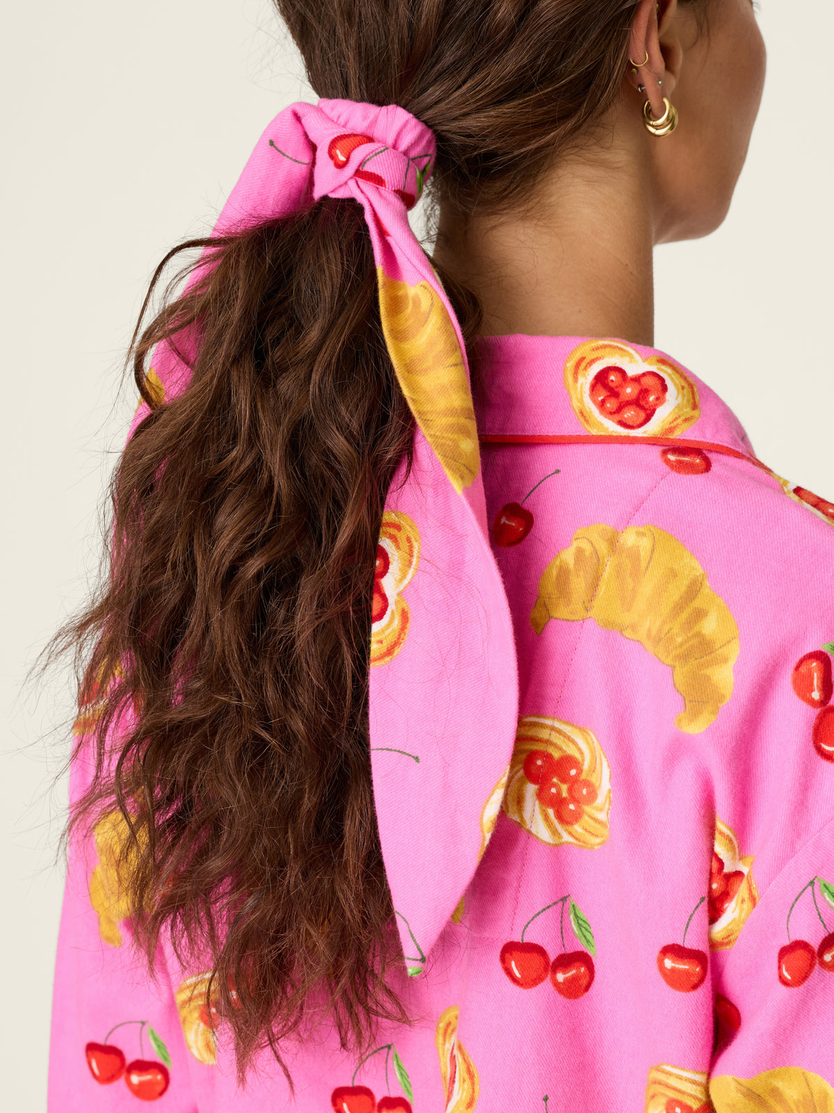 Long wavy brown hair tied with a pink and yellow scarf alongside a bright pink fruit-print top