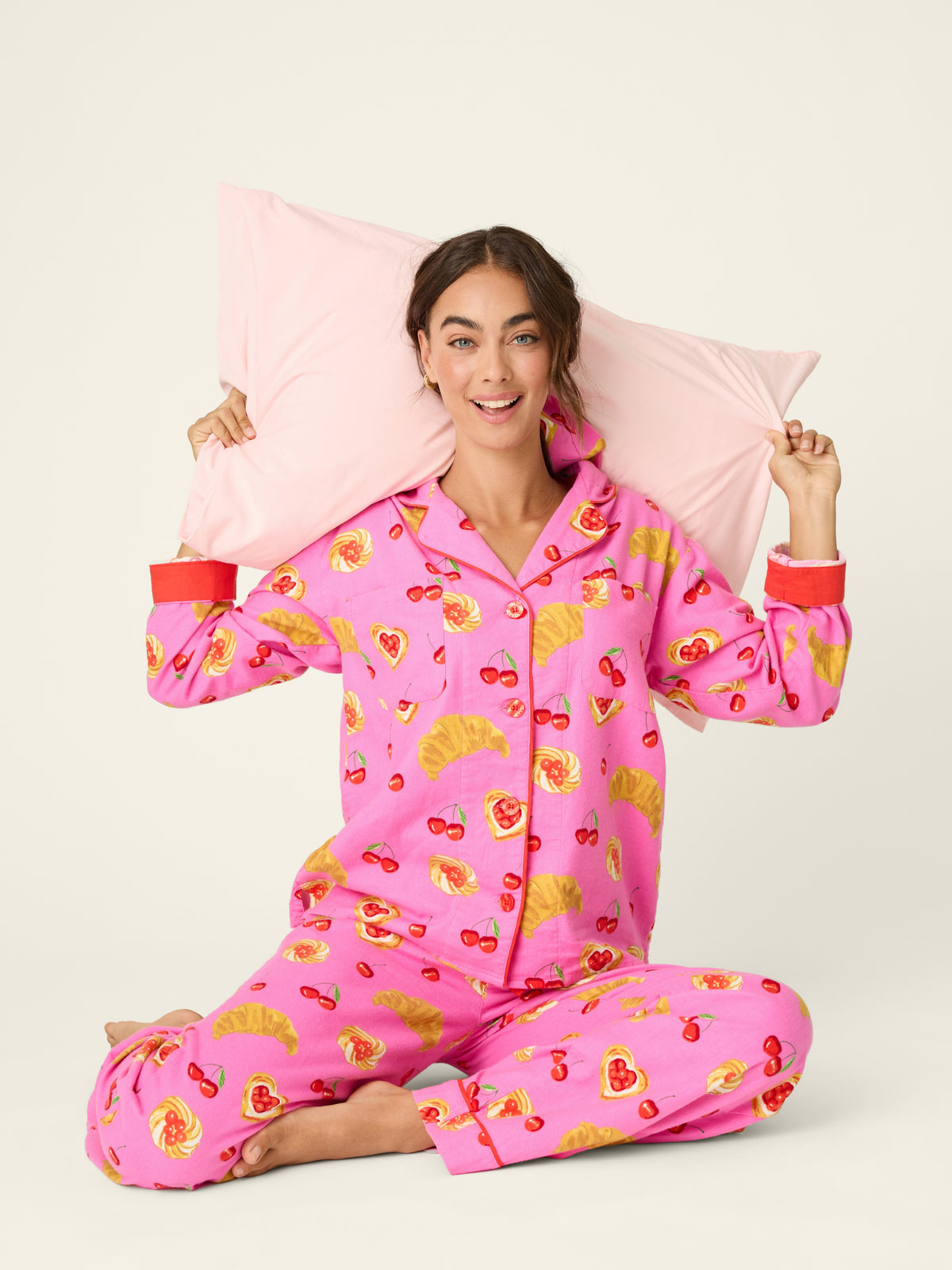 Model in pink printed pajamas sits cross-legged while holding a pale pink pillow behind her head