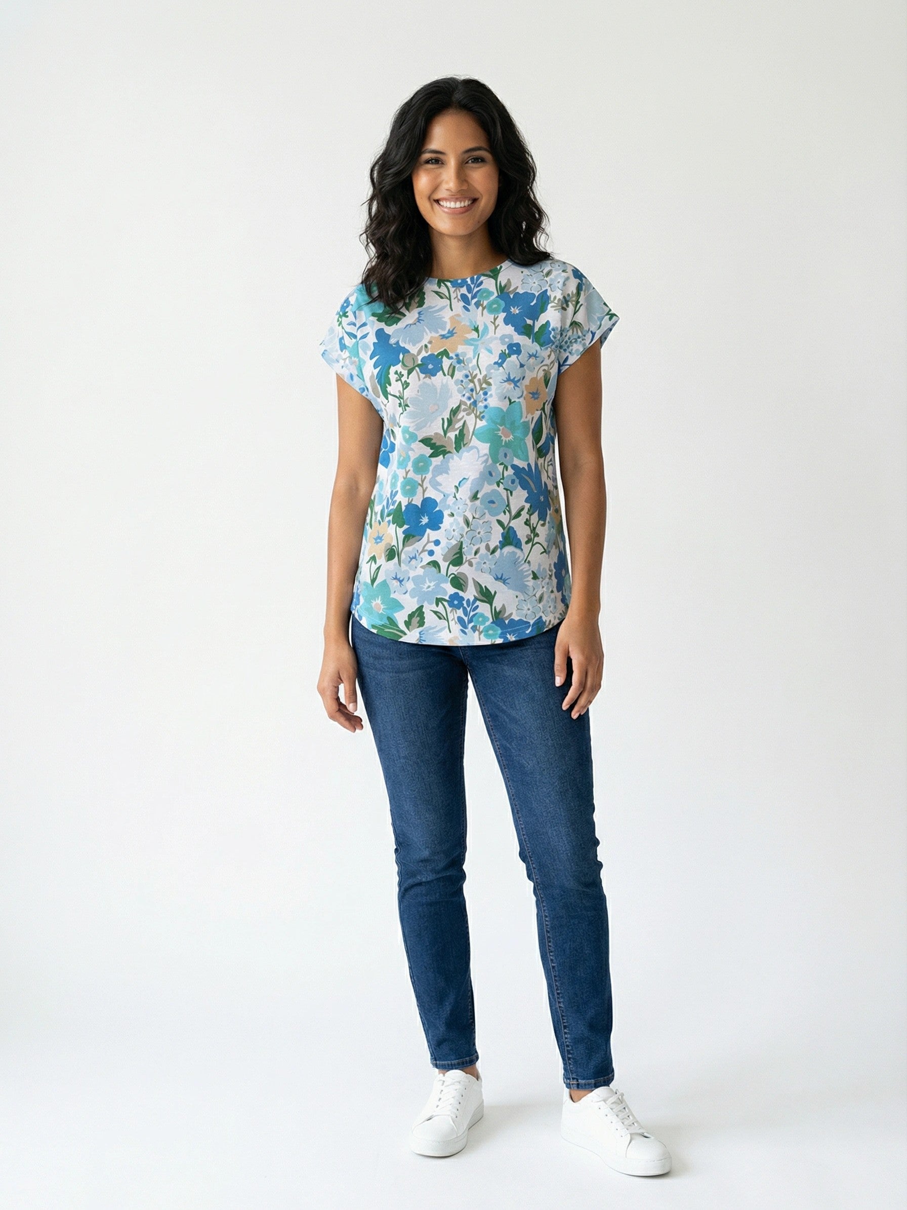 Person standing wearing a floral short-sleeve top with blue jeans and white sneakers on a plain white background