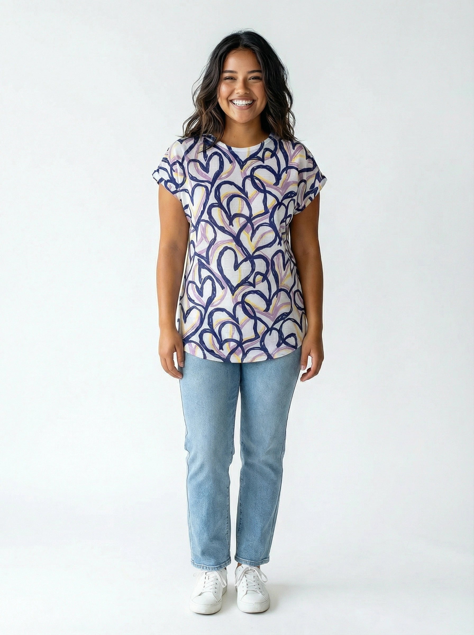 Smiling figure in a short-sleeve top with abstract heart patterns paired with light blue jeans and white sneakers.