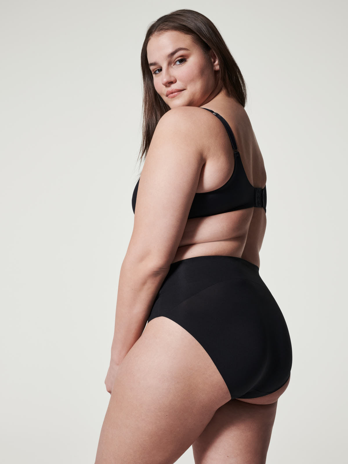 Curvy model wearing black SPANX Undie-Tectable® Brief by Spanx with a matching black bra against a neutral background