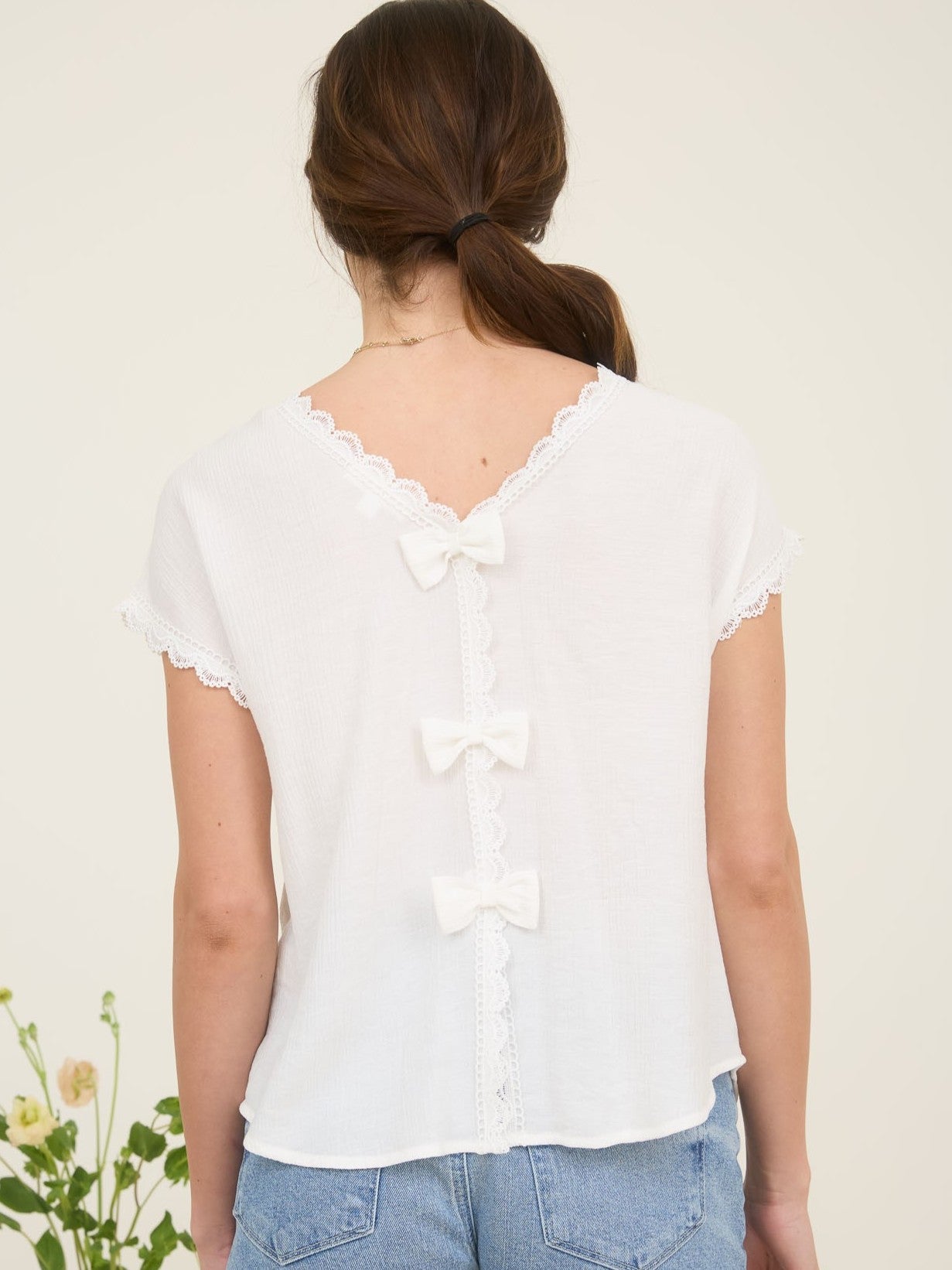 White short-sleeve top with lace trim and three decorative bows down the back, paired with blue denim jeans.