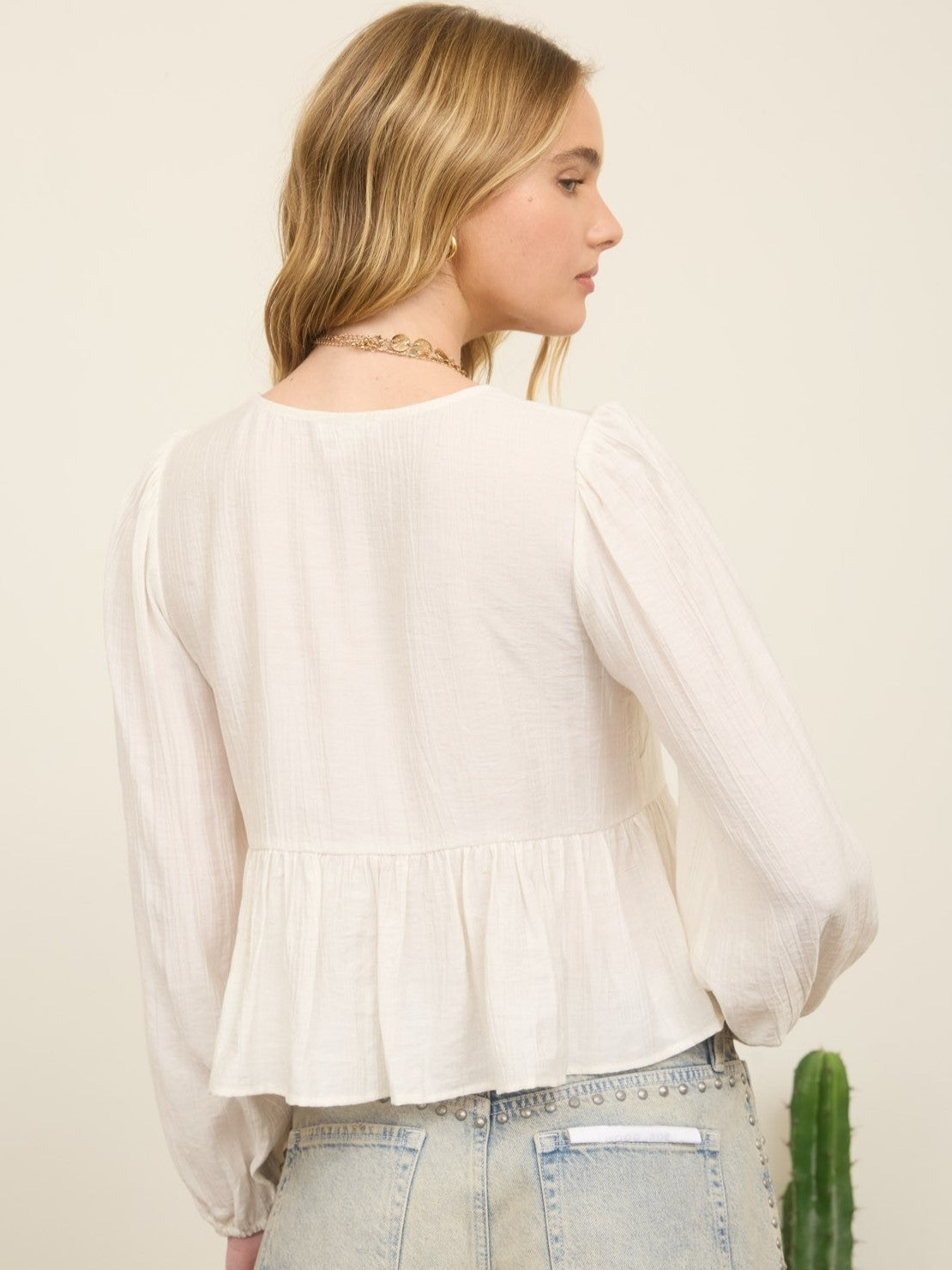 Back view of a white Floral Eyelet Front Tie Blouse by Mine with long sleeves and gathered hem paired with studded denim jeans
