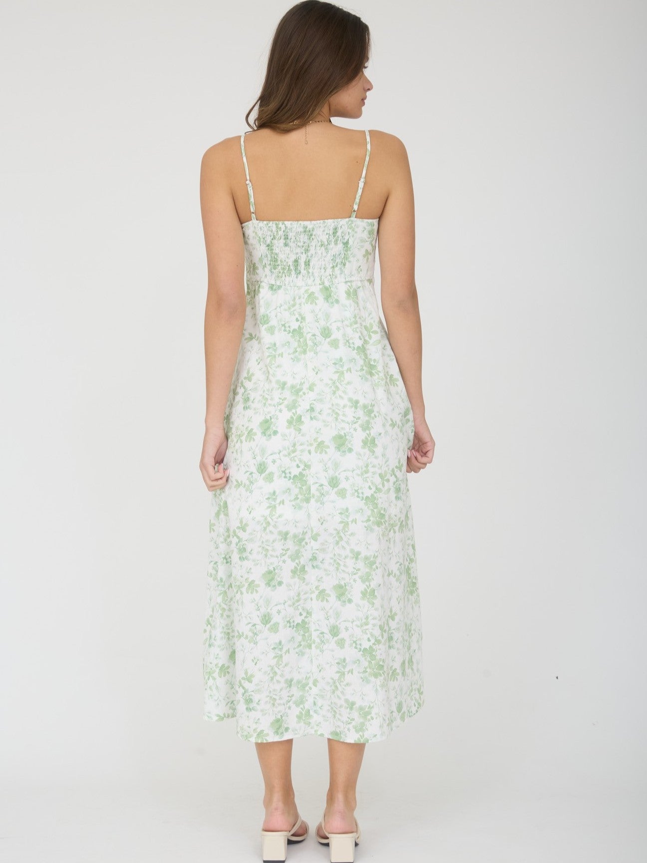 Back view of a white midi dress with green floral print and thin shoulder straps on a model standing against a plain background.
