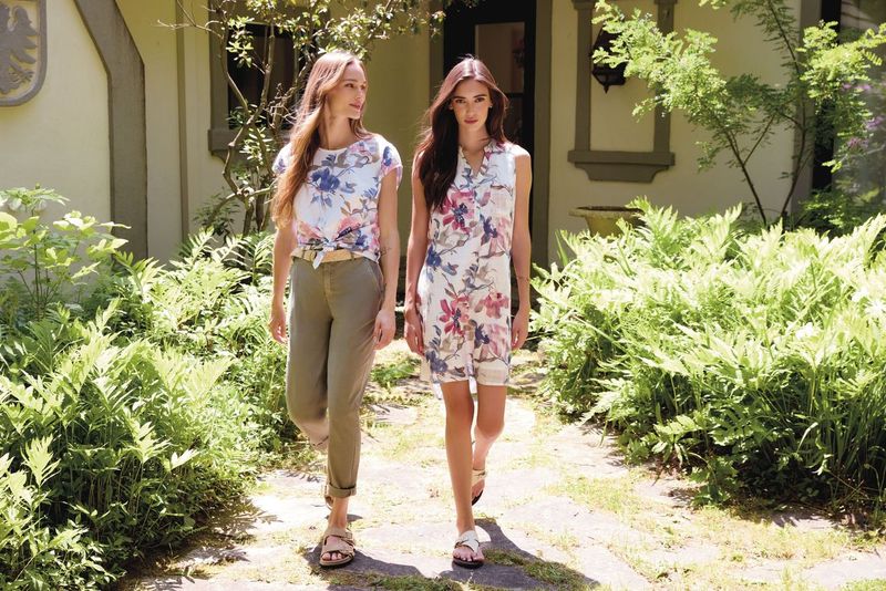 Two individuals walking outdoors on a sunlit path surrounded by green plants and trees near a building entrance