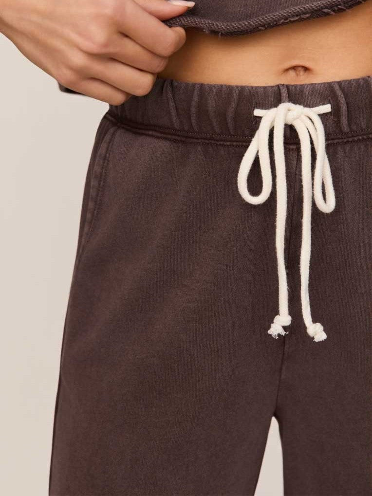 Close-up of dark brown sweatpants with white drawstring tied at the elastic waistband against a beige background.