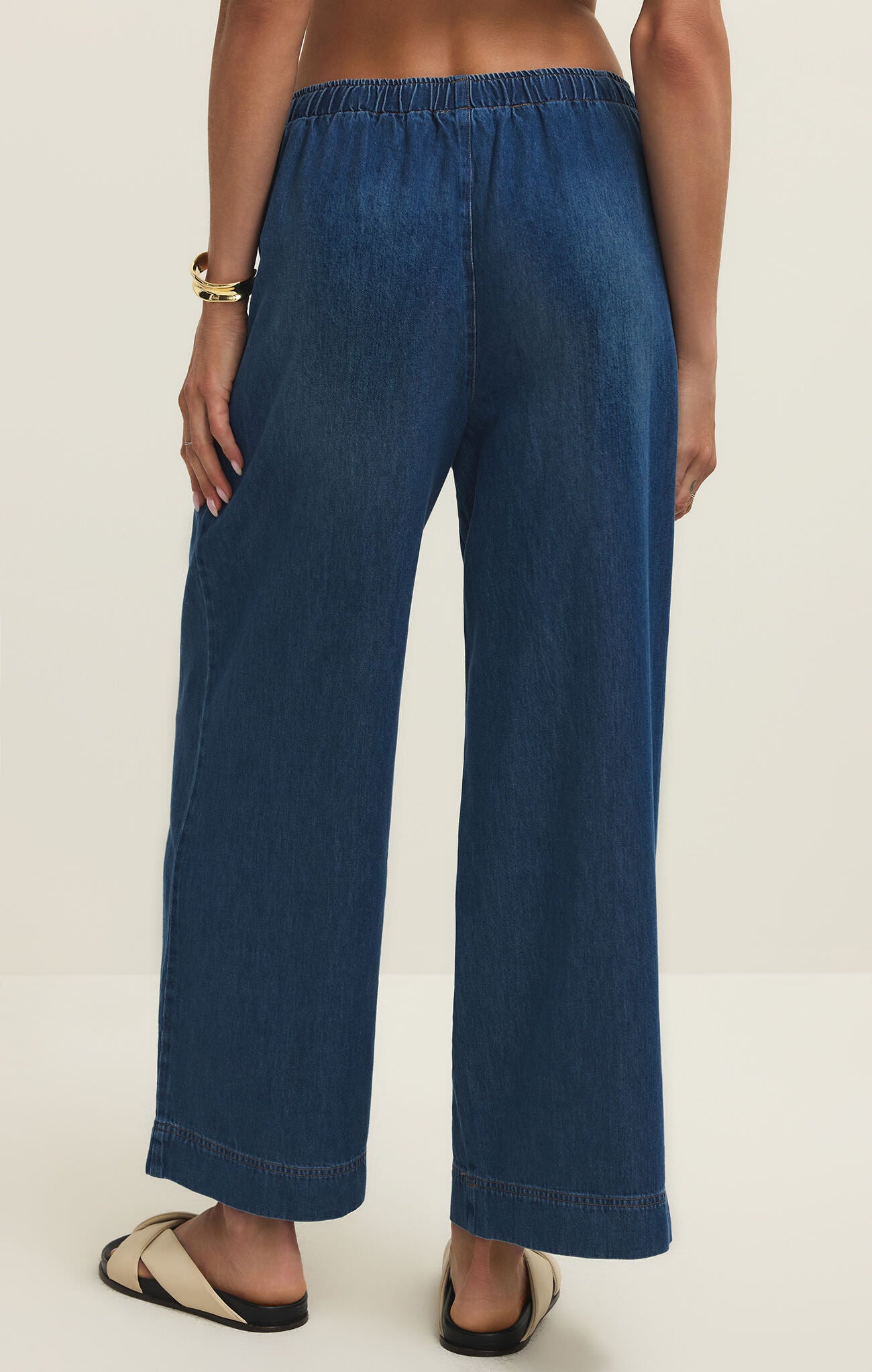 Back view of loose-fitting dark blue denim pants with elastic waistband worn with beige sandals and gold bracelet