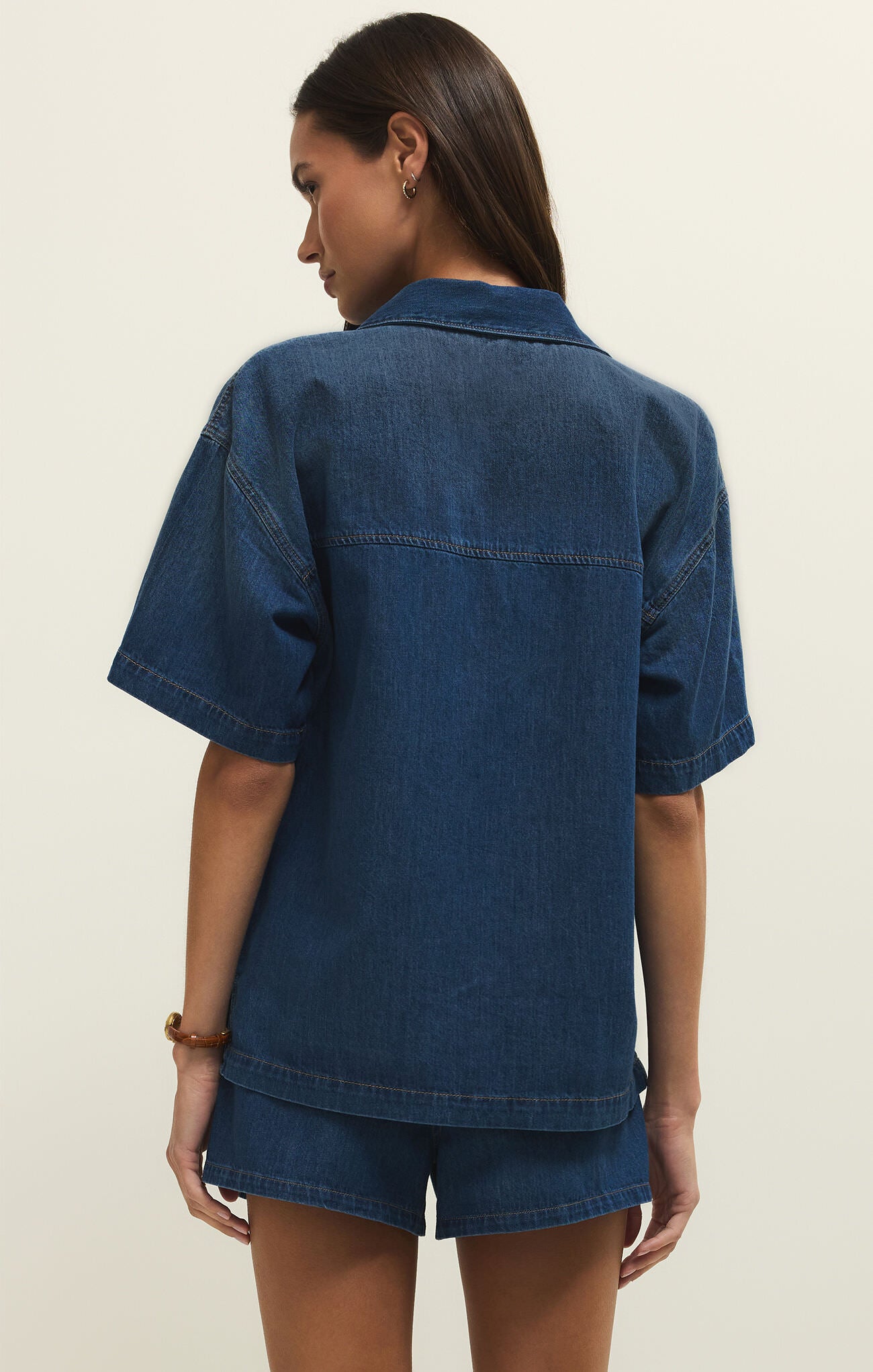 Back view of a person wearing dark blue denim short-sleeve jacket and matching denim shorts against a plain background