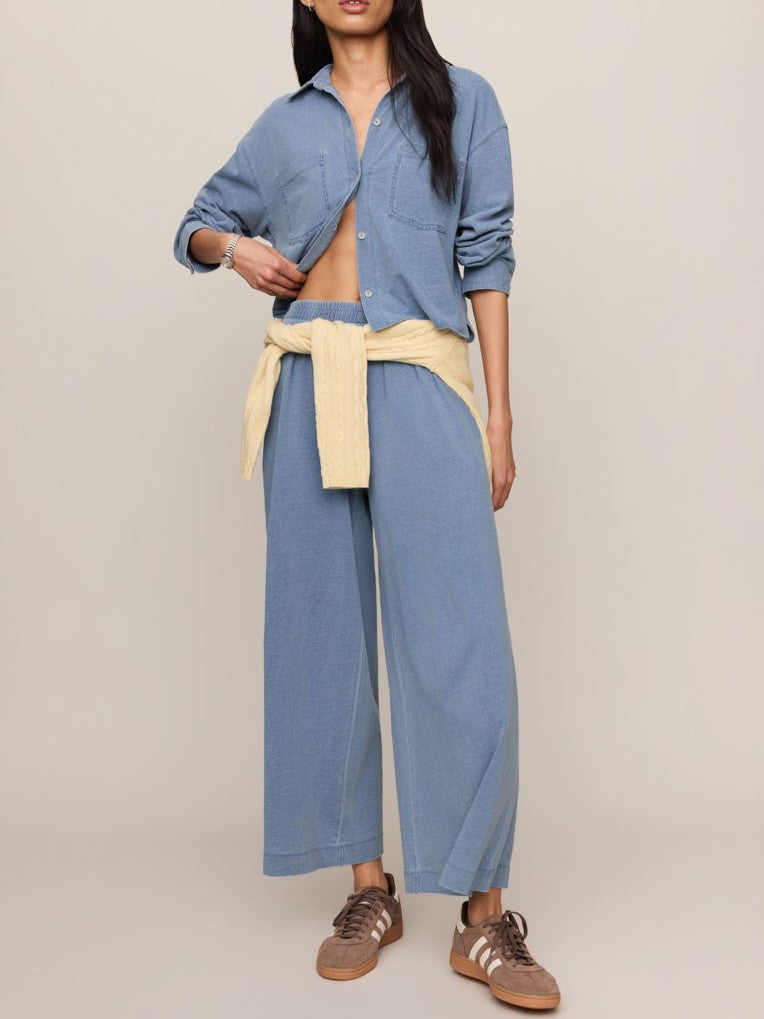 Loose-fitting blue button-up shirt and matching wide-leg pants with cream sweater tied around waist and brown sneakers