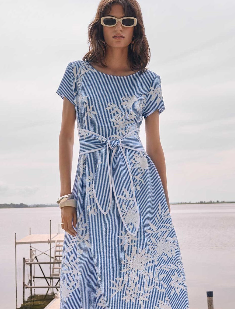 Blue and white striped dress with floral pattern and waist tie, paired with rectangular sunglasses and bracelets outdoors by water.